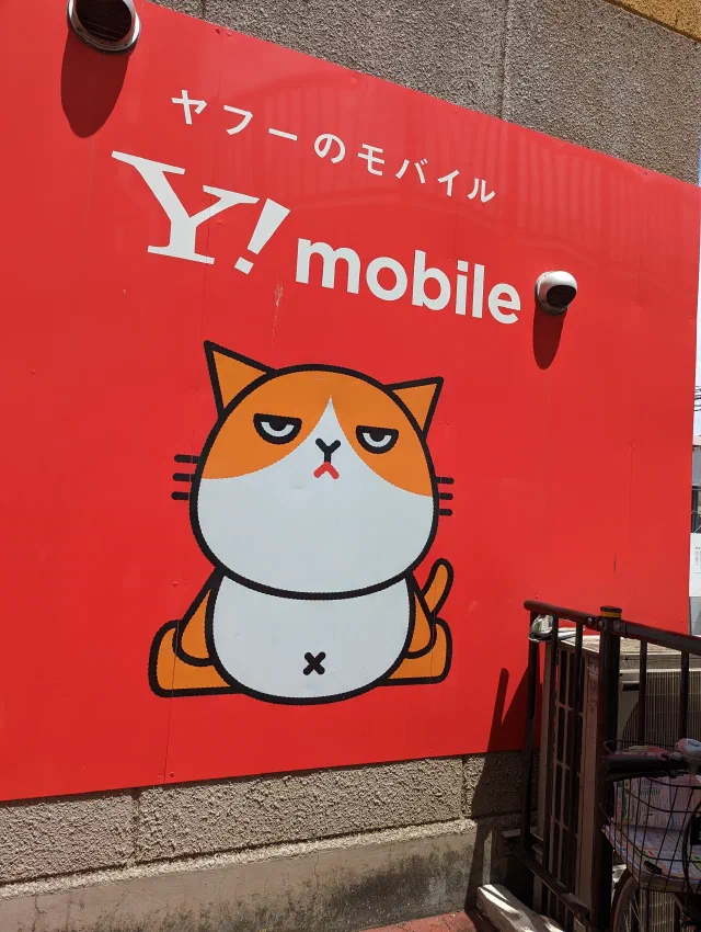 A cartoon orange and white cat with half-closed eyes and a frown, sitting on a red Y!mobile sign on a building wall, with a railing and a bicycle in the corner.