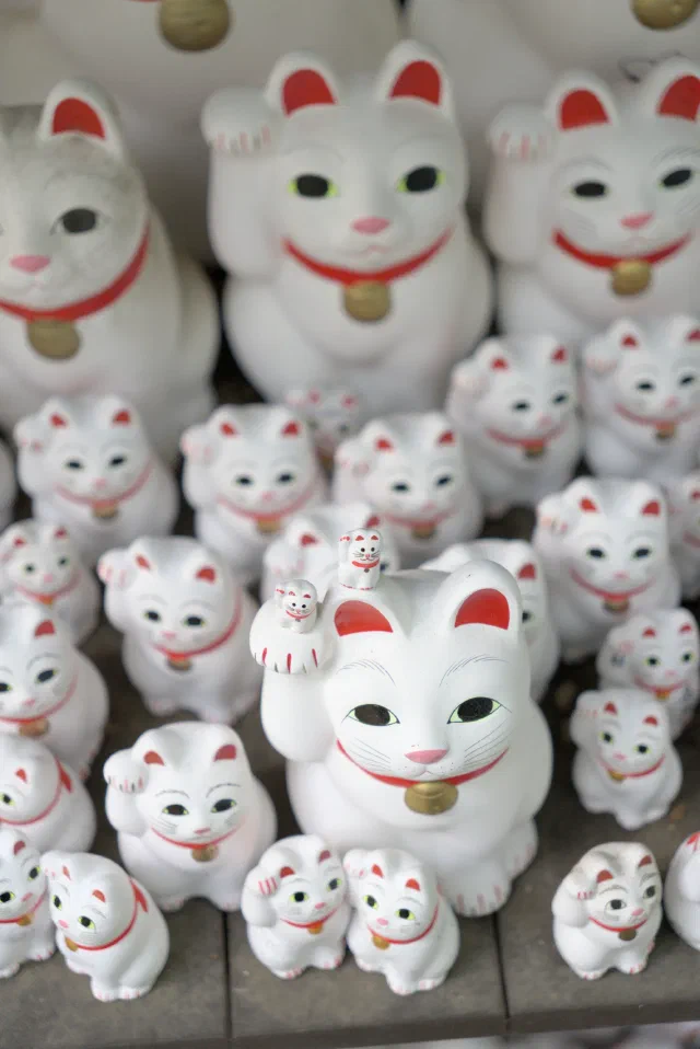 Rows of white maneki-neko figures with red ears, red collars and gold bells, each with a paw raised, crowded onto wooden shelves, with two tiny ones perched on the raised paw of a larger cat in the middle.