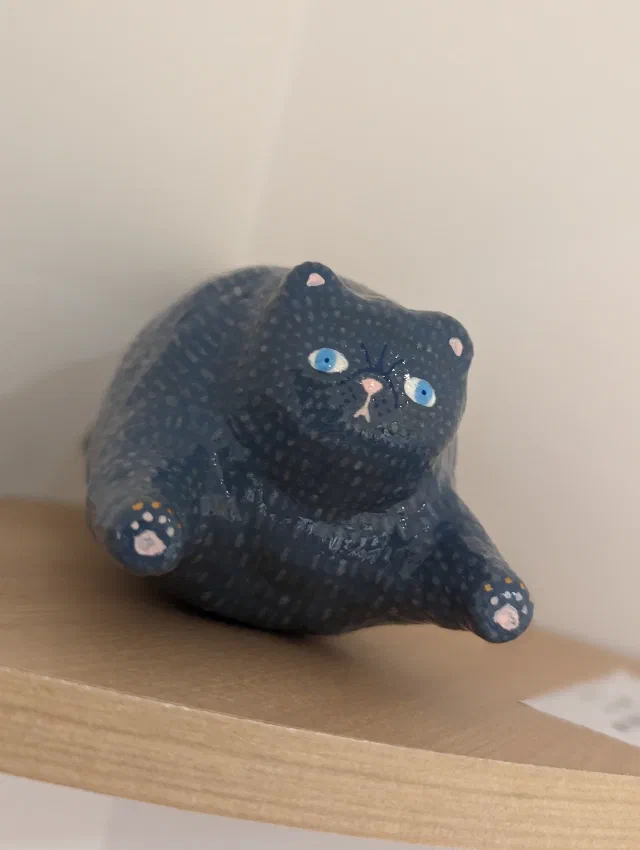 A handmade dark blue cat figure covered in tiny pale dots, with blue eyes, pink ears and nose and its front paws reaching out, lying on a curved wooden shelf.