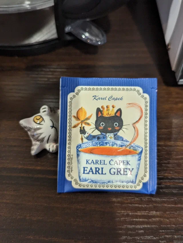 A Karel Čapek Earl Grey tea bag illustrated with a black cat in a crown holding a sceptre over a teacup, next to a small ceramic tabby-and-white cat lying on its back on a dark wooden table.