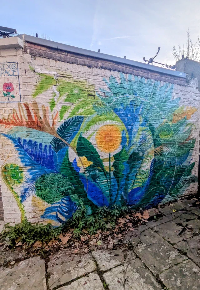 Ferns and leaves in blues and greens around an orange flower, painted on a whitewashed brick wall above a paved path.