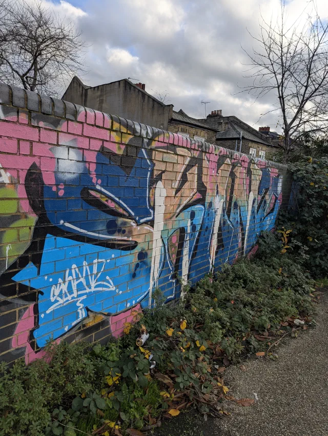 A long brick wall painted with large blue, pink and white graffiti letters, running along an overgrown path.