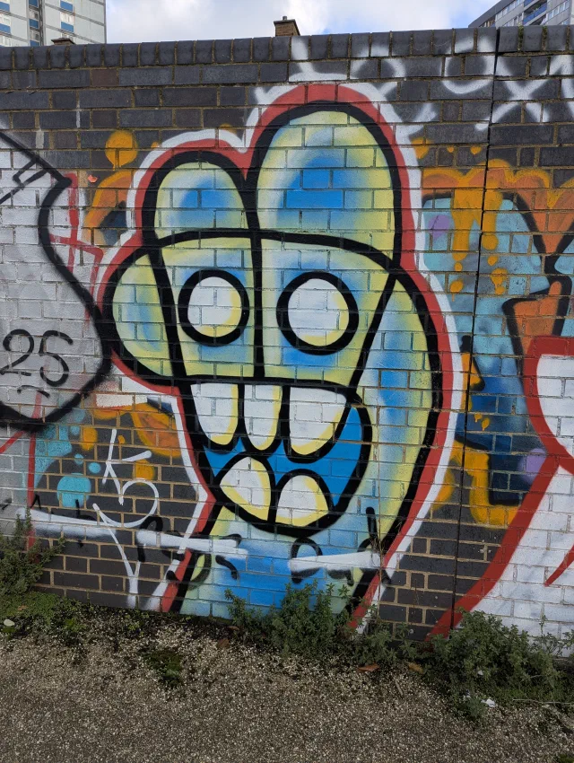 A yellow and blue cartoon face with round eyes and big square teeth, outlined in black and red on a brick wall.