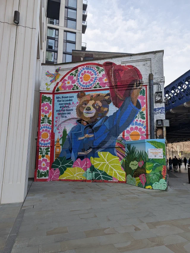 Mural of Paddington Bear in his blue duffle coat raising his red hat, framed by Andean flowers and leaves, with Big Ben and the London Eye behind him.