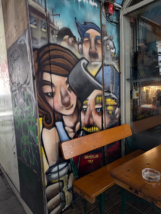 Painted faces of a woman with auburn hair and two men crowded together on a pillar, behind a wooden bench labelled Waterloo Tap.