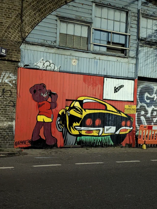 A maroon teddy bear in a red top drinking from a bottle beside a yellow muscle car, painted on a red wooden front under a railway arch.