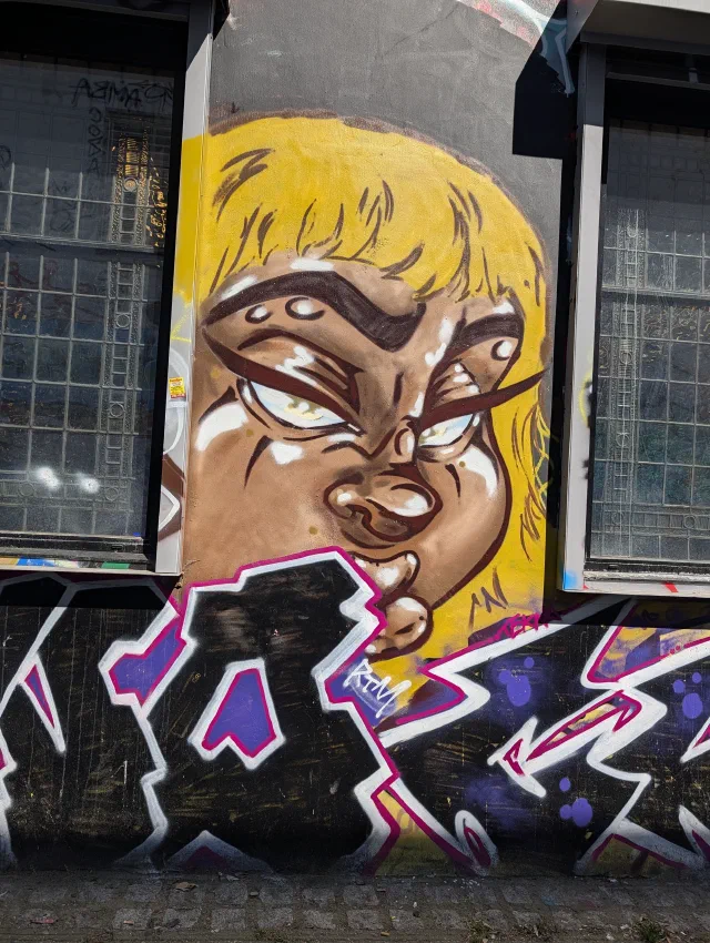 A cartoon face with yellow hair and a scowl giving a sideways look, painted between two windows above purple and pink graffiti letters.