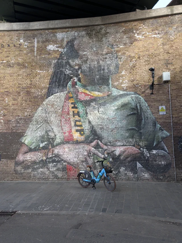 Large faded portrait of a bearded man in a green shirt and a red, yellow and green scarf, hands together, on a brick wall under a bridge with a hire bike parked in front.