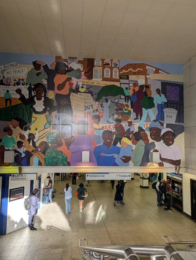 A huge painted mural of crowds at a market and a protest above the ticket hall of a tube station, with signs for the Victoria line below.