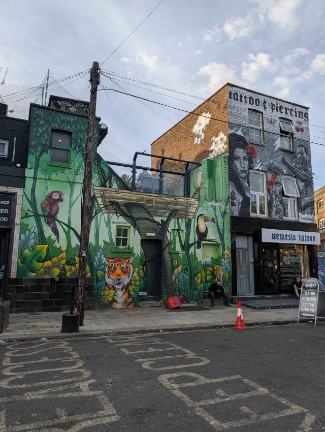 Two buildings covered in murals: a jungle with a parrot, toucan and tiger on the left, and a black and white woman with a red rose on a tattoo studio on the right.