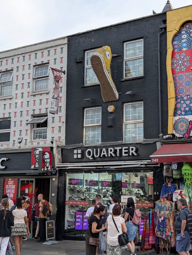 A giant yellow Converse trainer fixed to the front of a black shoe shop on Camden High Street, between a KFC and a building painted with mandalas.