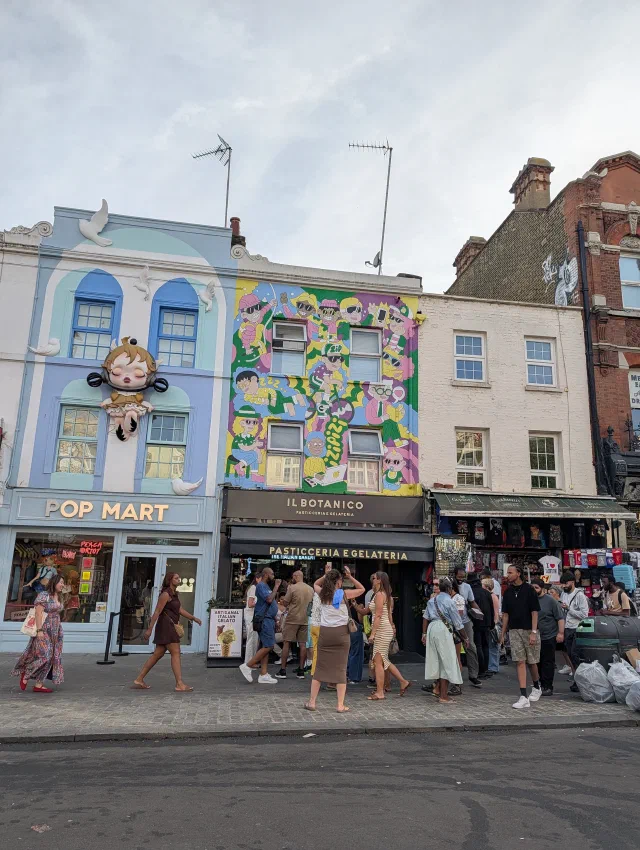A pale blue Pop Mart shopfront with a giant doll above the door, beside a café painted with rows of cartoon characters in pink, yellow and green.