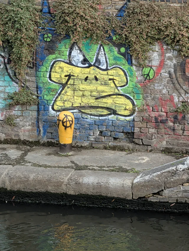 A yellow cartoon duck face with a grumpy beak on a canal wall, behind a yellow bollard with an anarchy symbol.