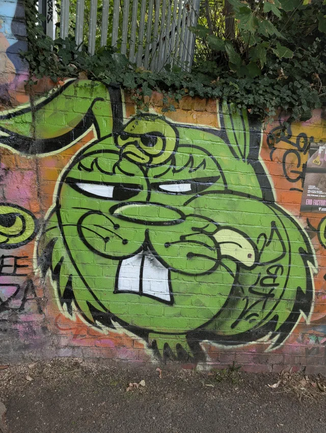 A grumpy green cartoon rabbit with big front teeth on an orange and pink wall, under ivy and railings.