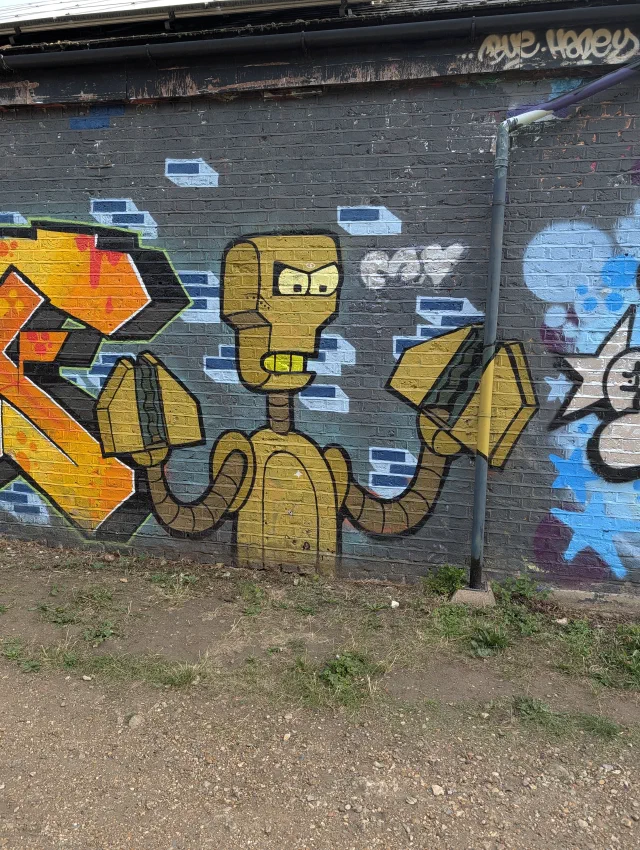A yellow cartoon robot in the style of The Simpsons, with bendy arms holding up two blocks, on a grey brick wall.