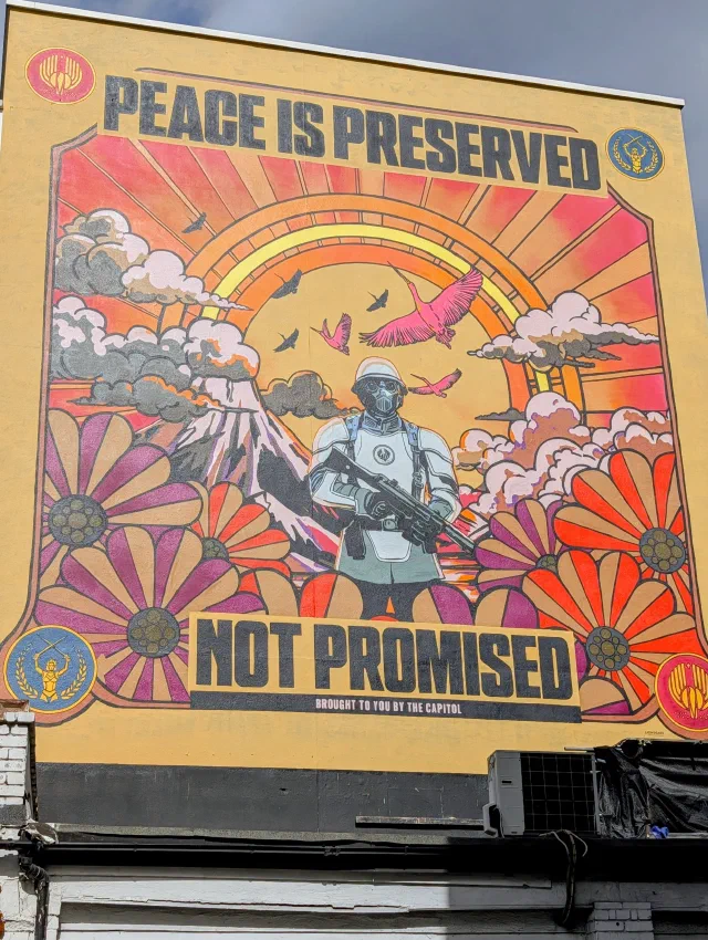 Propaganda-style poster mural of a white-armoured soldier with a rifle among giant flowers, a volcano and a rainbow, reading Peace Is Preserved Not Promised.