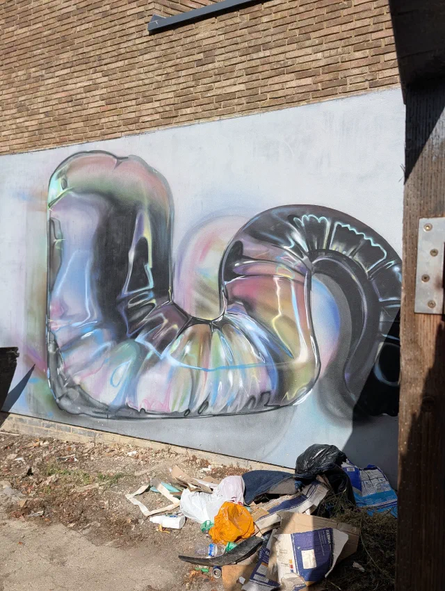 A shiny chrome letter shape reflecting pinks and blues, painted on a pale grey wall above a pile of rubbish.