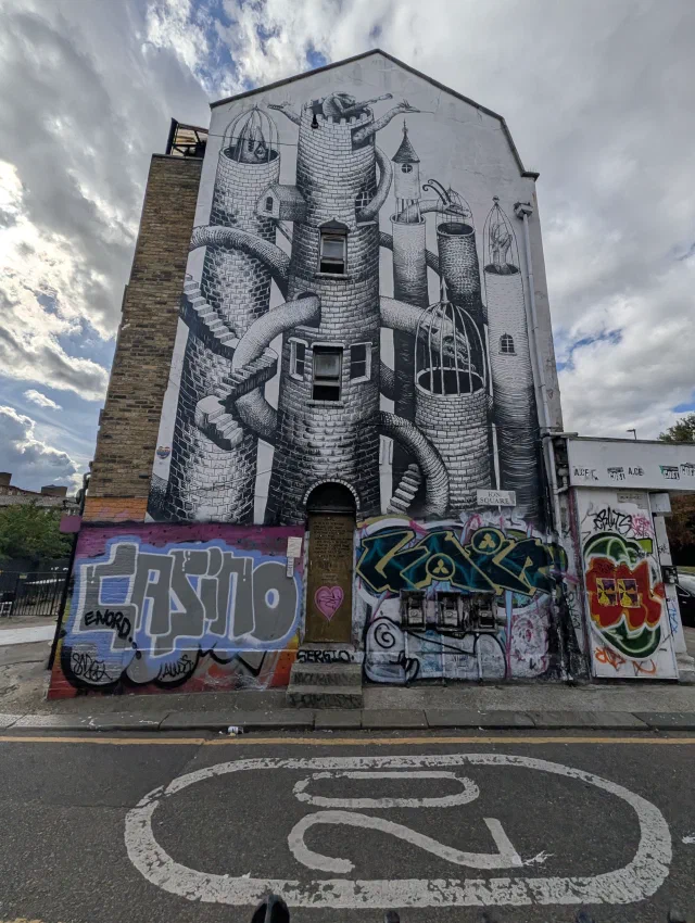Black and white mural filling the gable end of a building, of brick towers linked by pipes and staircases with birdcages on top, above graffiti on the ground floor.