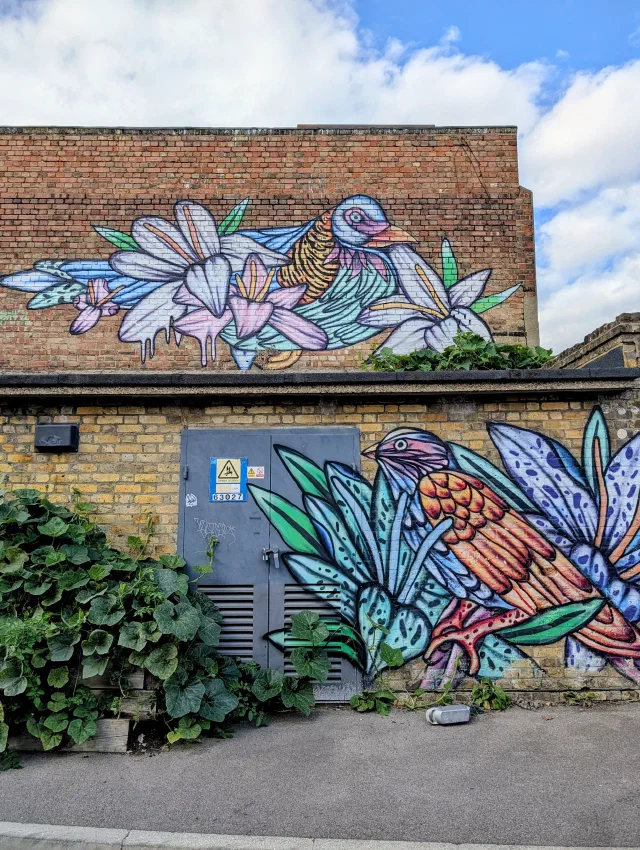 Two colourful birds among white lilies and blue leaves, one painted along the top of a brick building and one beside a grey substation door.