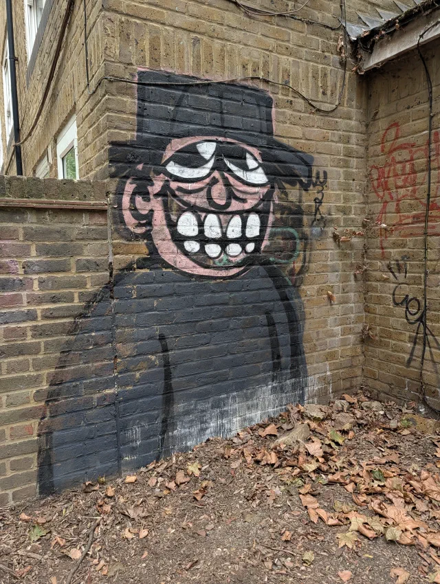 A grinning pink-faced character in a black top hat and cloak, painted in the corner of a brick yard.