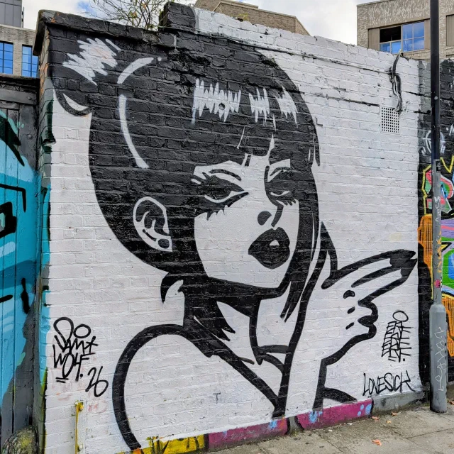 Black and white comic-style woman with a bob, pointing a finger gun, on a white wall.