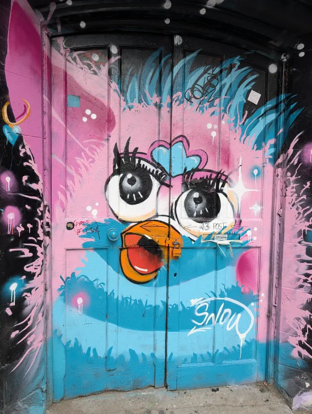 A pink and blue Furby with huge eyes and an orange beak, painted over a pair of old double doors.