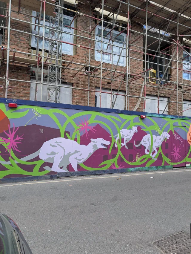 Pale greyhounds racing across a purple and green hoarding of twisting vines, in front of a building under scaffolding.