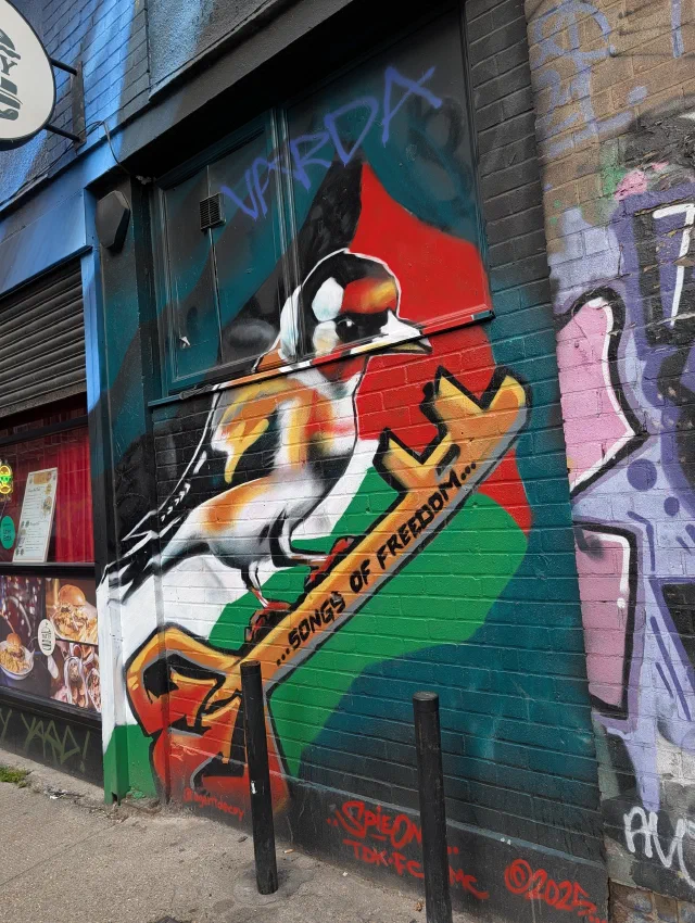 A goldfinch perched on a branch with the words Songs of freedom, painted over the colours of the Palestinian flag on the corner of a building.