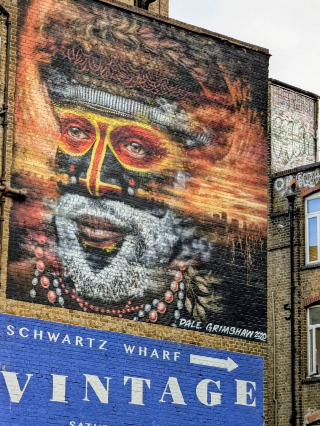 A large portrait of a man with red and yellow face paint, a feathered headdress and a white beard, high on a wall above a Schwartz Wharf Vintage sign.