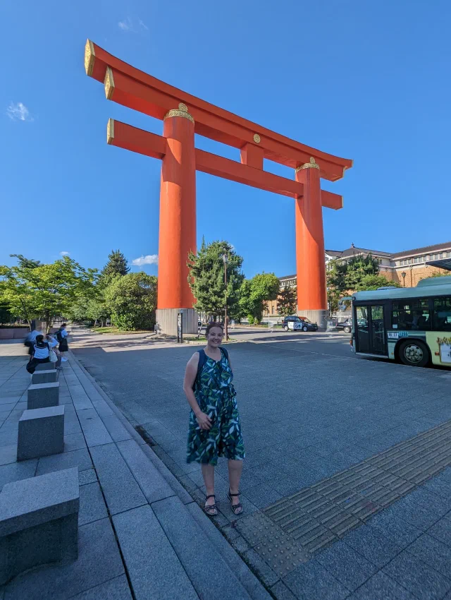 A huge vermilion torii with gold caps on its pillars rising over a wide paved square under a deep blue sky, with a smiling woman in a green patterned dress in the foreground and a bus passing on the right.