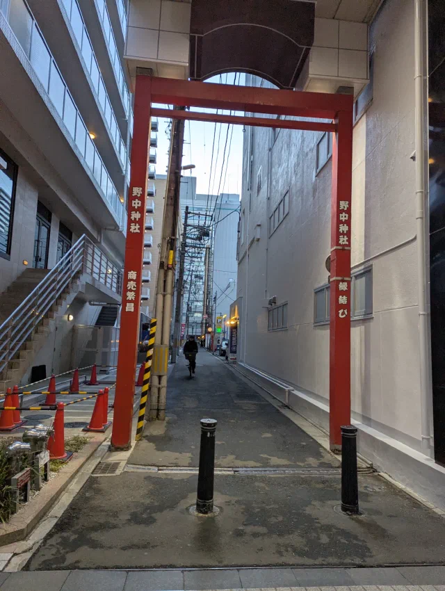 A tall, plain red gate painted with the shrine's name standing over a narrow side street between modern buildings, with a cyclist riding towards it and bollards in front.
