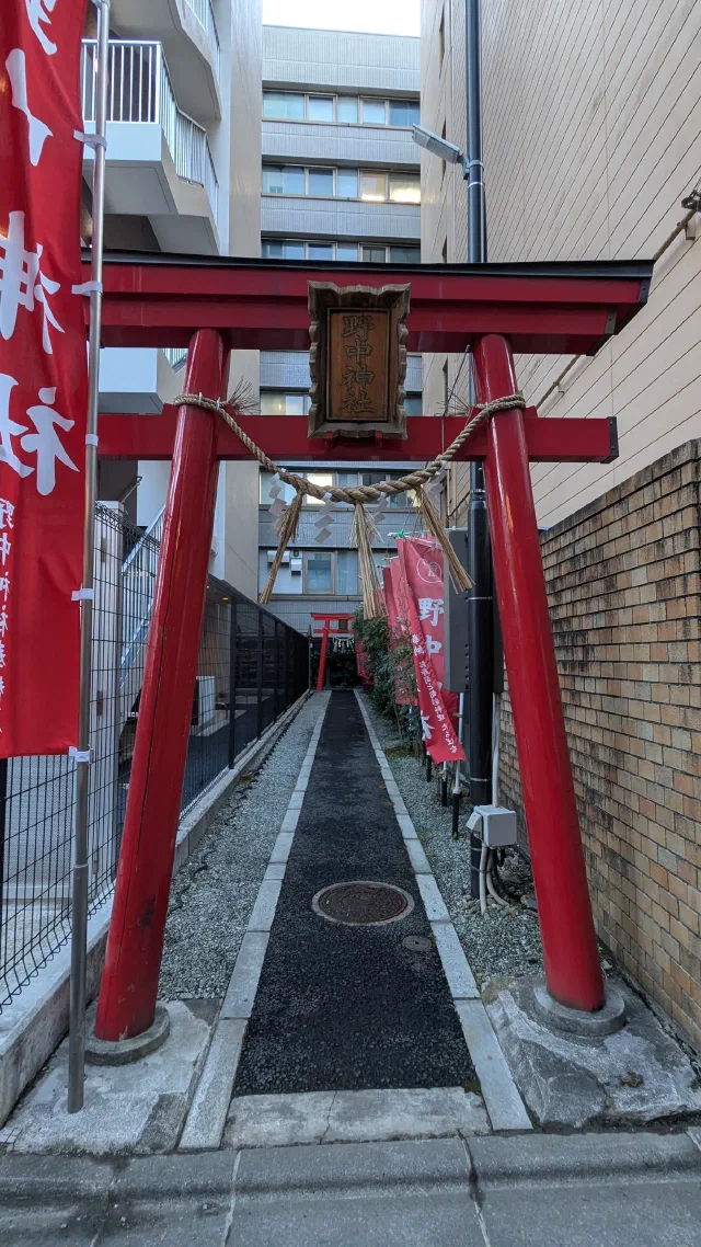 A red torii with a straw rope and a carved wooden plaque, framing a narrow path between apartment blocks, with red banners and a second small torii at the far end.