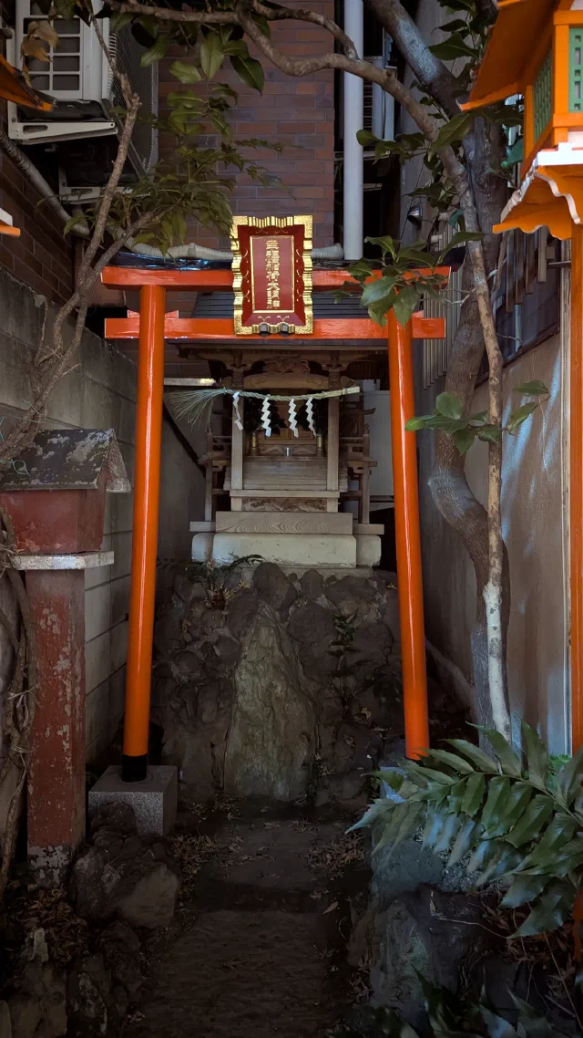 A small orange torii with a gold-framed red plaque in front of a tiny wooden shrine on a rock, squeezed between a brick wall and a building, with paper streamers and a palm leaf in the foreground.