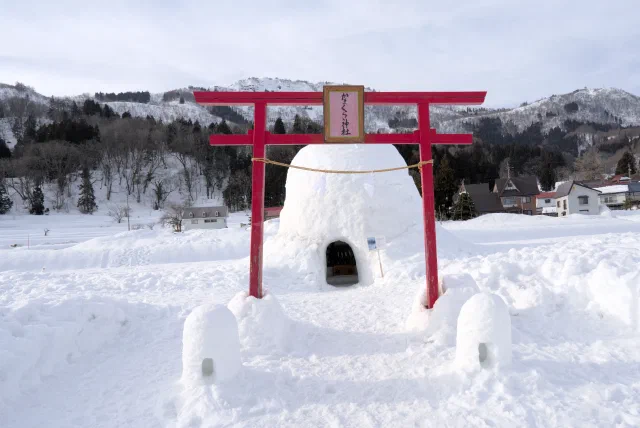 A red torii with a pink plaque in front of a domed snow hut with a small altar inside its doorway, flanked by two little snow lanterns, with snowy hills and houses behind.