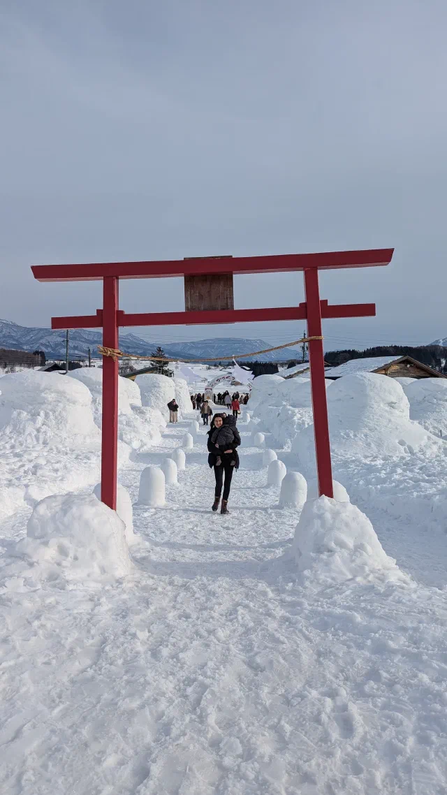 A red torii with a straw rope standing in deep snow at the start of a path lined with small snow lanterns and big snow mounds, with a woman walking towards the camera and mountains behind.