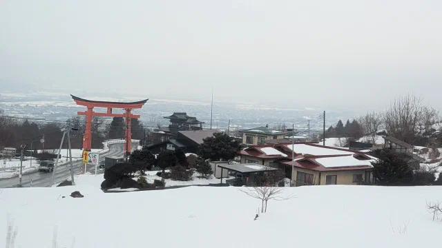 A big red torii beside a road on a snowy hillside, with snow-covered houses and a temple-style roof below it and a hazy, snowy plain stretching into the distance.