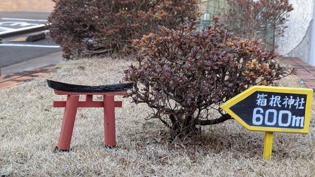 A small homemade red torii with a black top beam standing on frosty grass beside a shrub, next to a yellow arrow sign reading 箱根神社 600m.