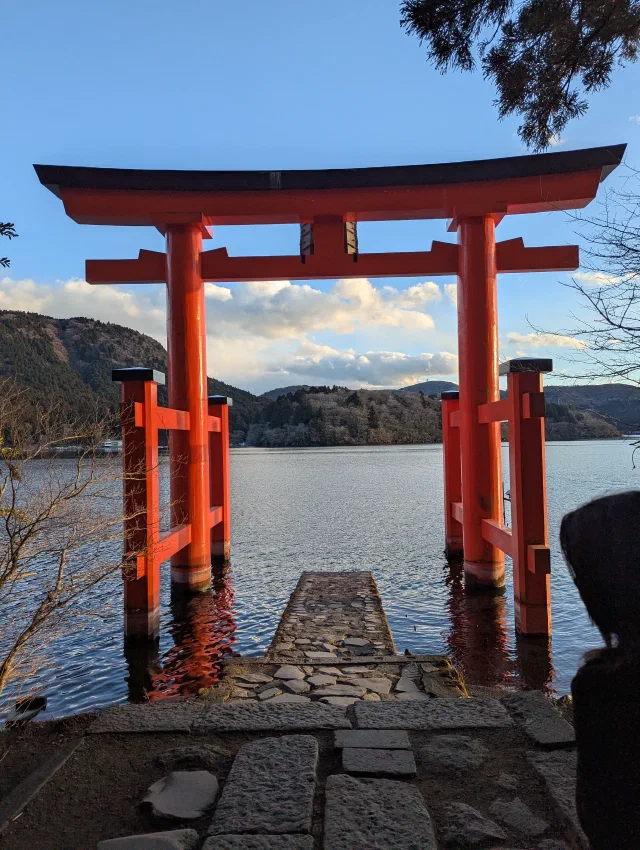 A red torii standing in the water at the end of a short stone jetty, with Lake Ashi, wooded hills and white clouds behind, and someone's hooded head at the right edge.
