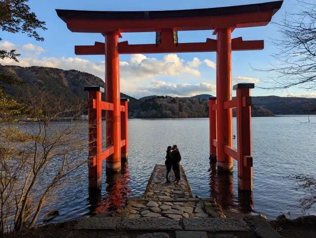 Two people embracing on the stone jetty between the pillars of the red torii, silhouetted against Lake Ashi, with wooded hills and clouds lit by low sun.