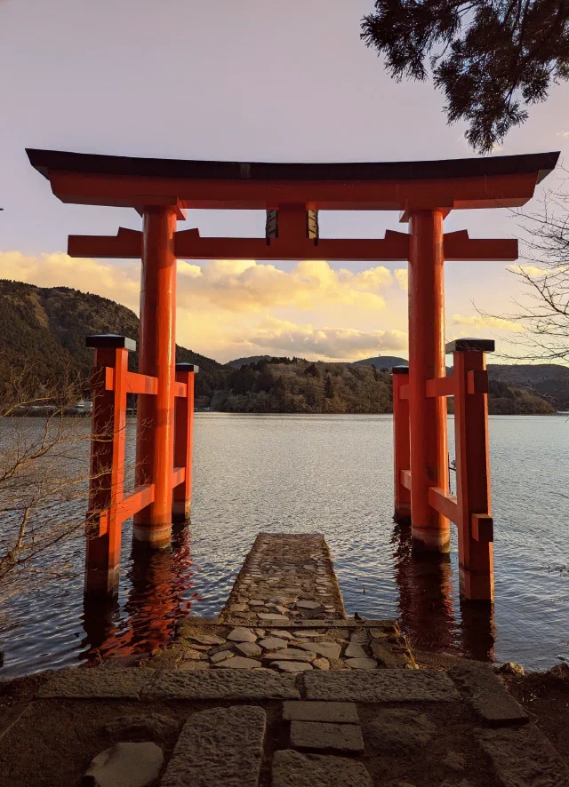 A red torii standing in the water at the end of a short stone jetty, lit by low golden sun, with Lake Ashi, wooded hills and glowing clouds behind.
