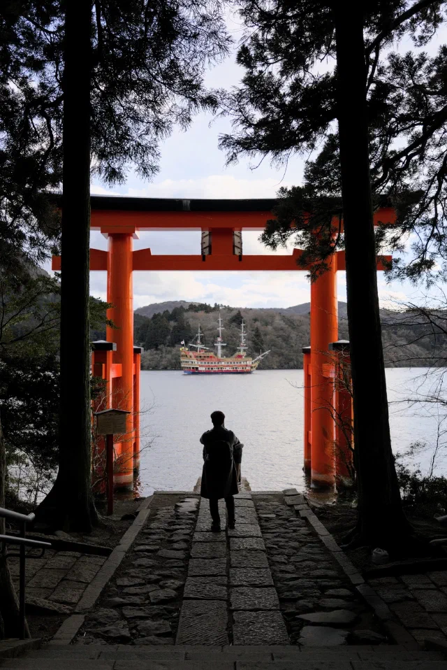 A person silhouetted on a stone path between tall cedar trunks, looking through a big red torii to Lake Ashi, where a replica sailing ship is passing.