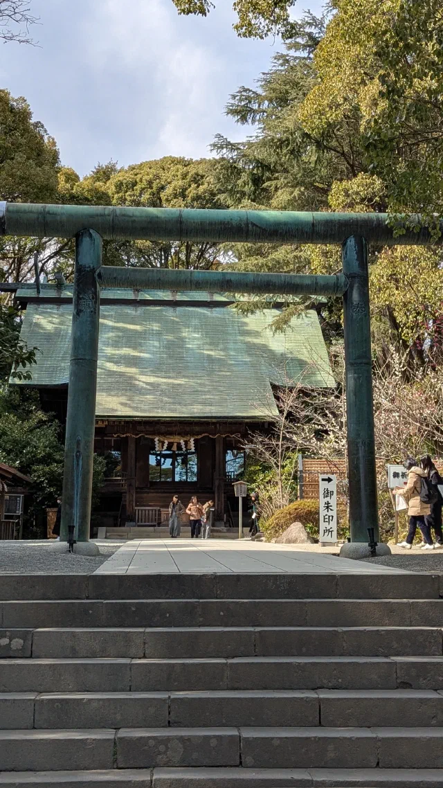 A green bronze torii at the top of stone steps, framing a shrine hall with a green copper roof and people walking in front.