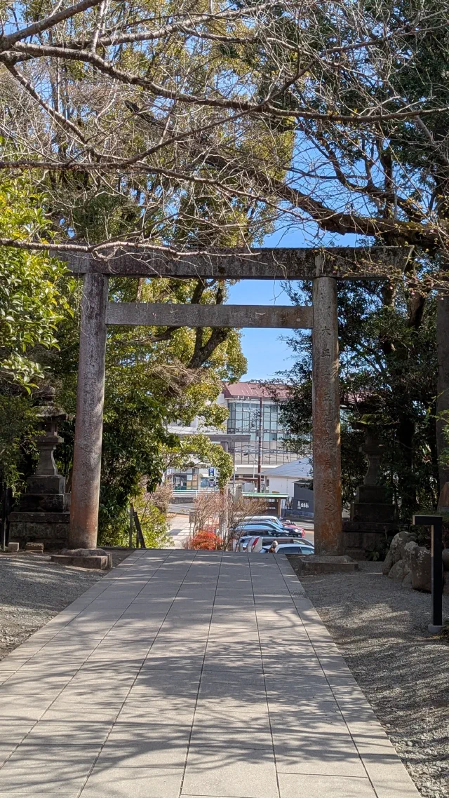 A weathered grey stone torii carved with inscriptions, at the end of a paved path under trees, with a car park and buildings beyond.