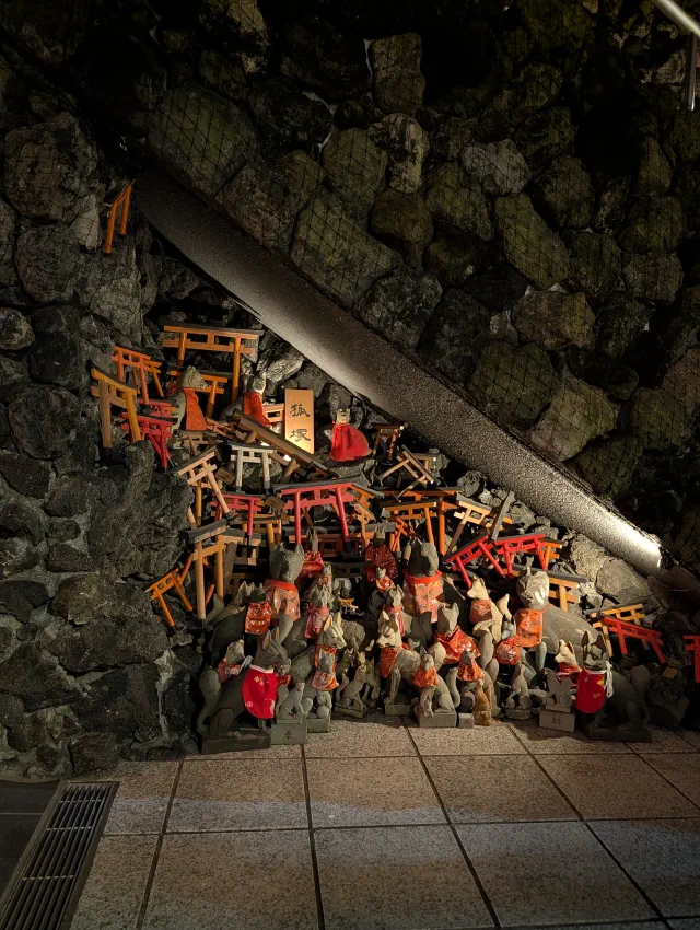 Dozens of small stone fox statues in red bibs crowded in front of a heap of miniature red and orange torii stacked against a mossy stone wall, lit at night.