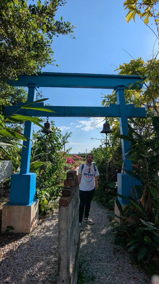 A bright blue torii with a bell hanging from each pillar over a gravel path through tropical plants, with a man in an Orion beer T-shirt standing underneath.