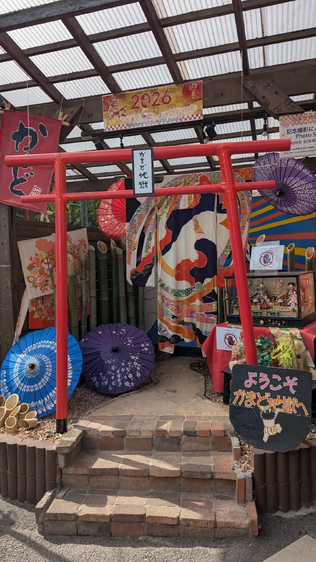 A red torii labelled かまど地獄 in a decorated photo spot under a glass roof, surrounded by paper parasols, banners and a 2026 sign, with a board reading ようこそかまど地獄へ.