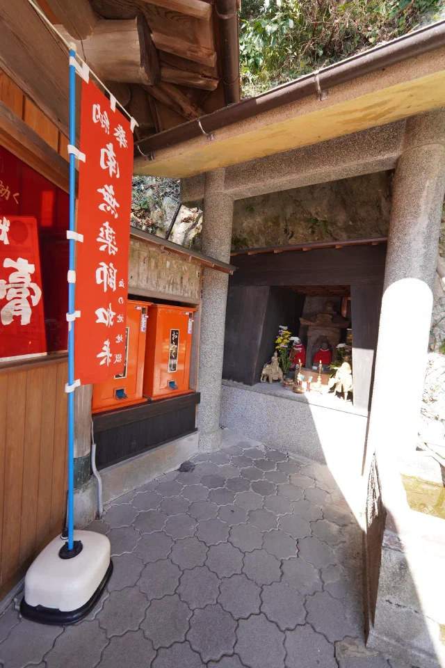 A small shrine in a rock niche framed by granite pillars under a wooden eave, with a statue, flowers and offerings inside and a red banner on the left.