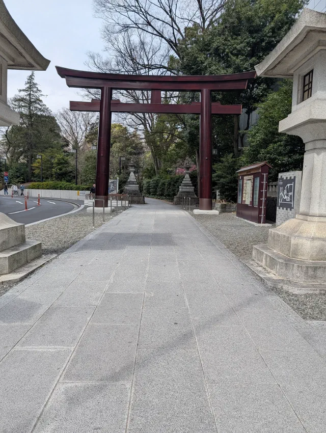 A dark red torii over a wide paved approach lined with stone lanterns and trees, with a road curving past on the left.