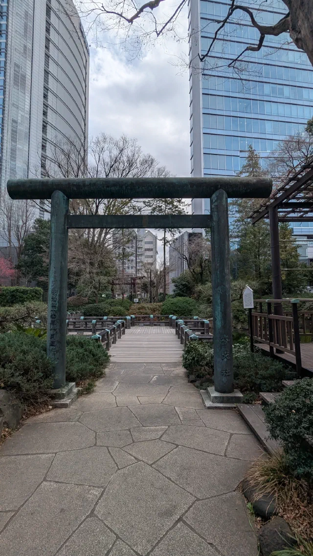 A green bronze torii at the start of a wooden walkway through a garden, with tall glass office towers rising behind bare trees.
