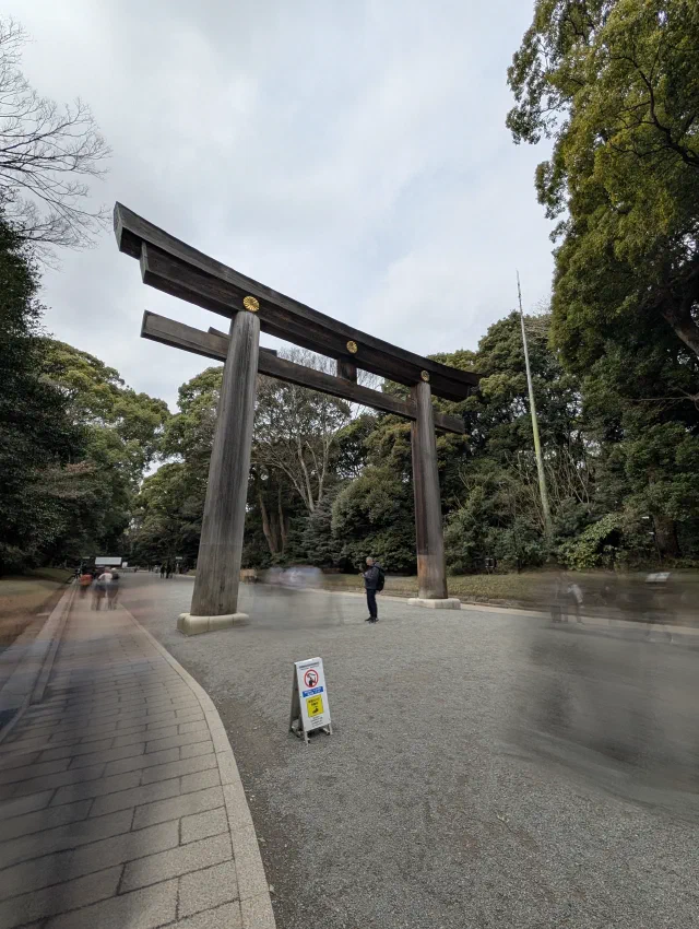 A huge plain wooden torii with gold chrysanthemum crests over a gravel path through woods, with the people walking under it blurred into ghostly streaks and one person standing still.